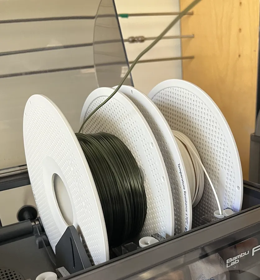 Three filament spools seated side by side in the AMS bays, with dark green filament feeding over the top of the roll