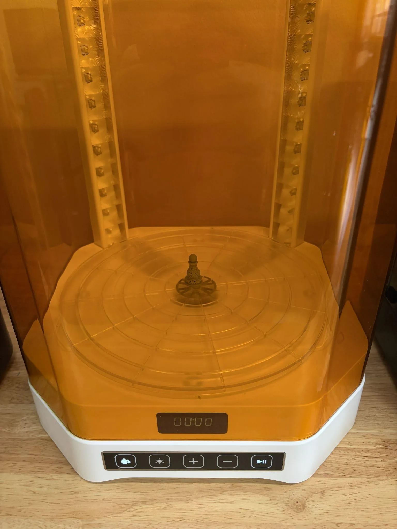 A grey chess pawn standing in the centre of the curing turntable under the UV lamps, seen through the orange cover