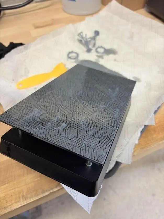The build plate wiped clean and lying face-up on a bench beside a plastic scraper and used cleaning pieces