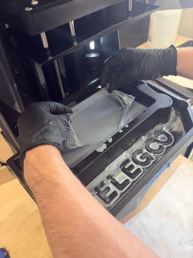 Two gloved hands easing the cured resin sheet up off the bottom of the tank by the two stems