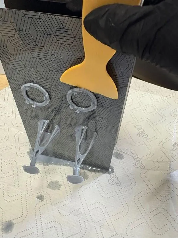 A gloved hand using a yellow plastic scraper to lift cured resin pieces off the build plate