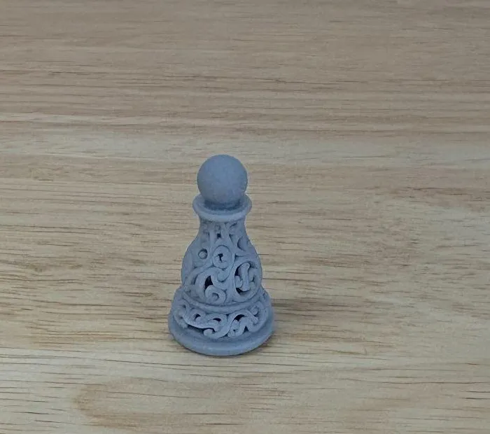 A small grey resin chess pawn with fine scrollwork over its whole surface, standing on a workbench