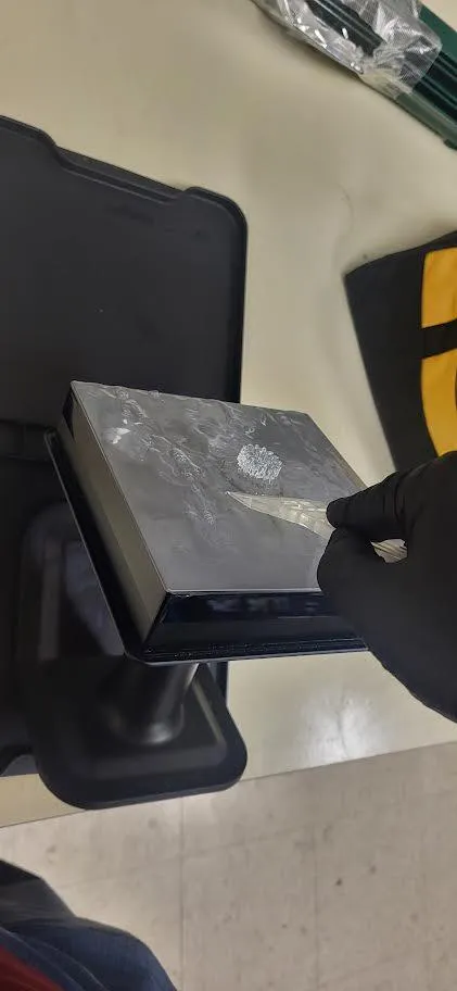 A gloved hand easing a plastic scraper under the edge of a print's raft on the angled build platform