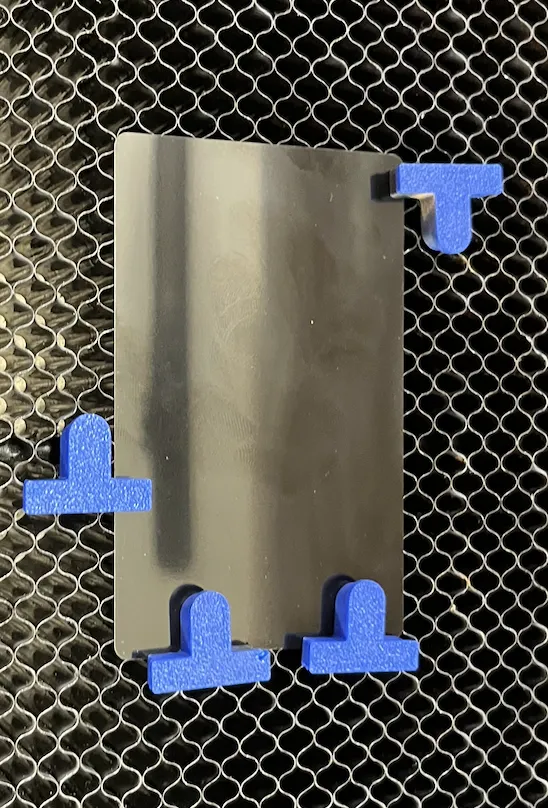 A dark metallic business card blank sitting on the Glowforge honeycomb bed, held in place by four blue T-shaped hold-down pins along its edges