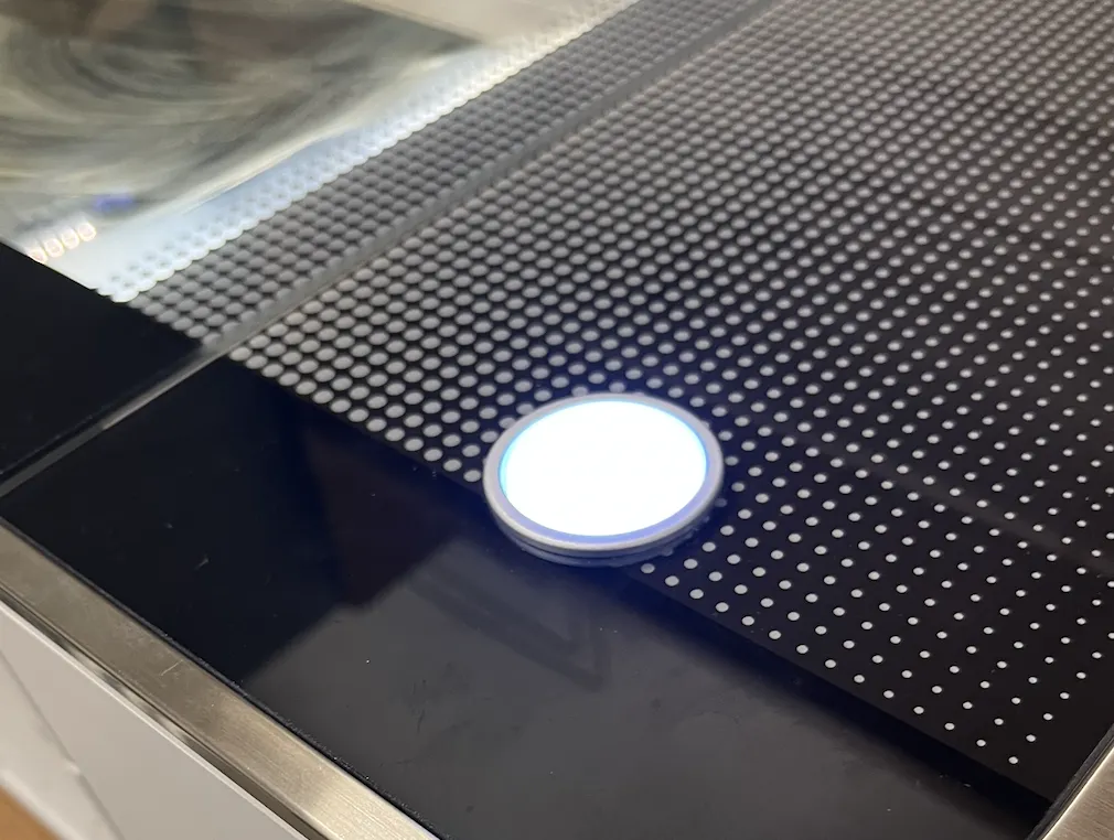 The Glowforge's round start button glowing bright white against the dark machine bed