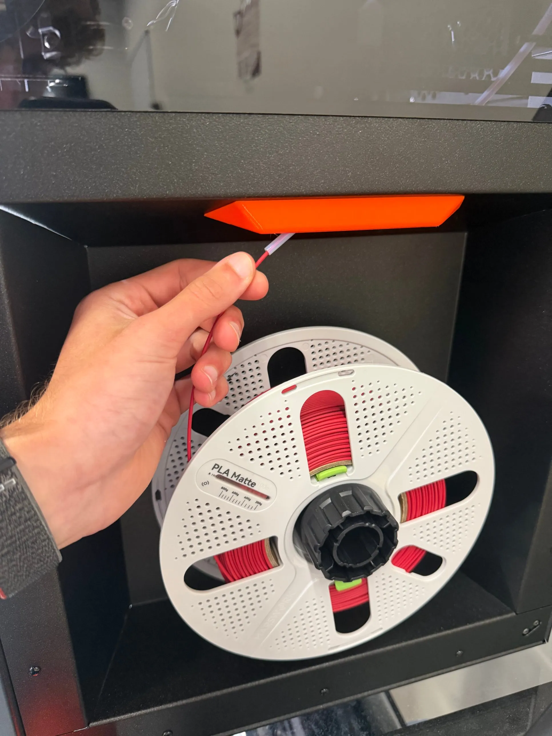 A hand holding the filament just outside the white PTFE tube, with the spool on its holder below